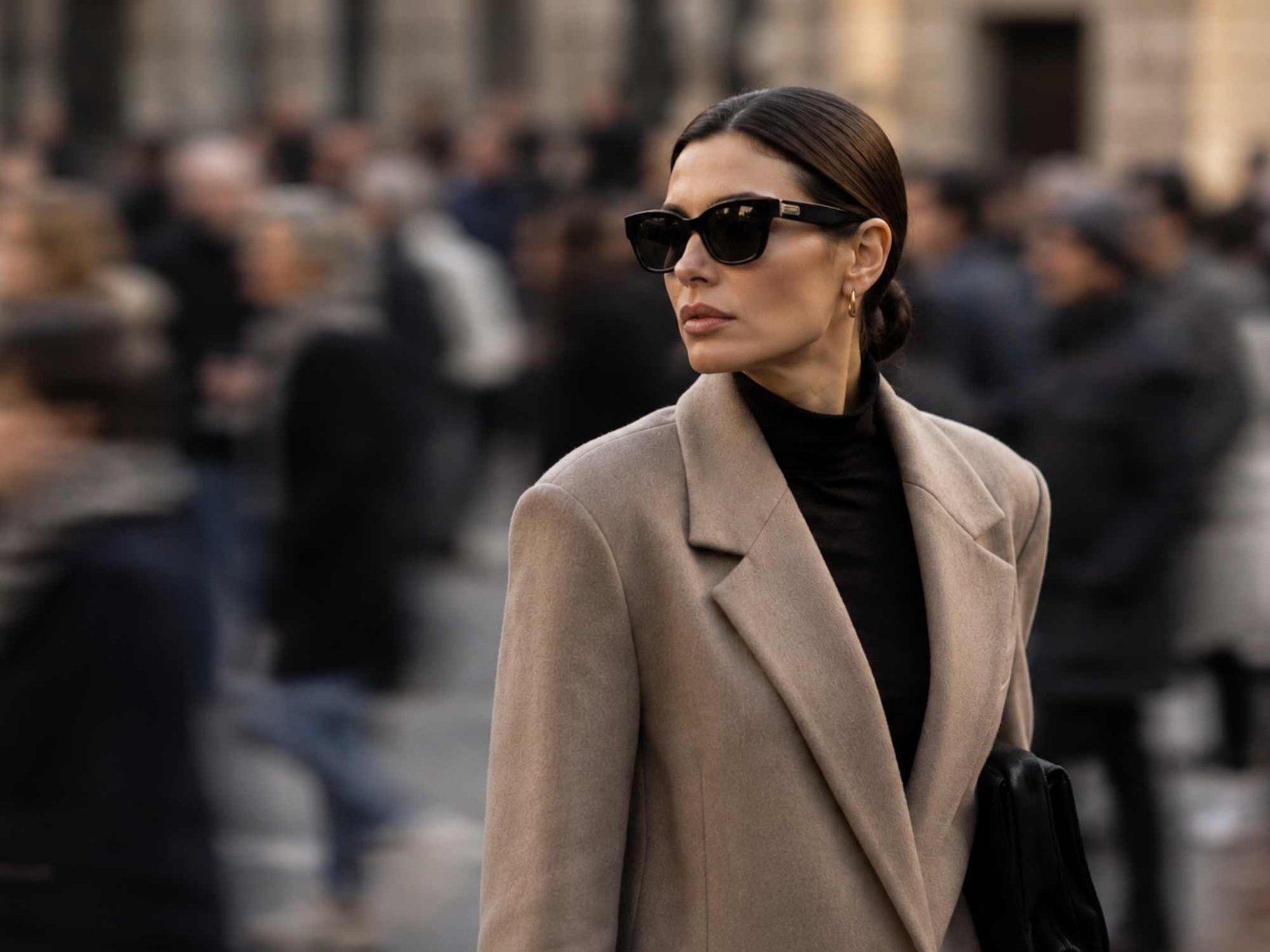 Una donna con un cappotto cammello, maglione nero e occhiali da Sole in mezzo alla folla