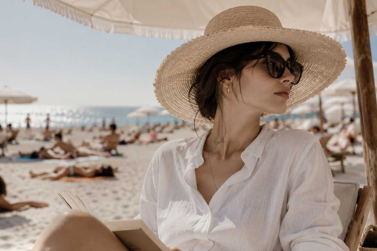 una ragazza si trova sotto un ombrellone e indossa un cappello di paglia e una camicia oversize di lino
